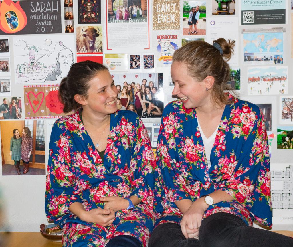 Mara Steinitz ’18 and Sarah Kolenbrander ’18 sport matching bridesmaid robes in front of “The Wall” of memories in their living room. Photo by Lorna Sherwood Caballero ’18.